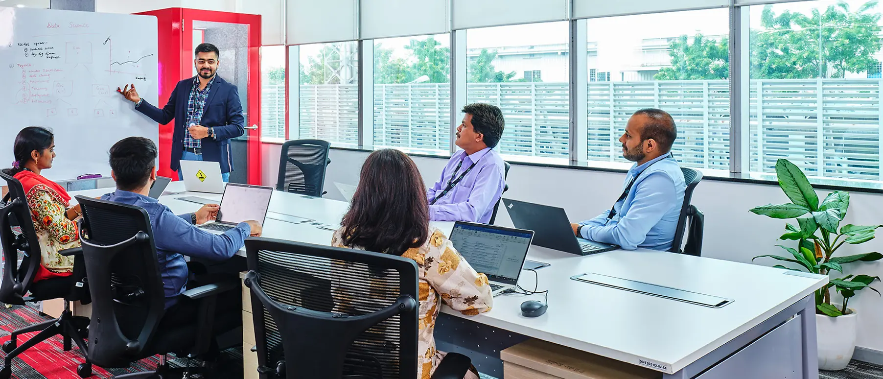 Five men and women are gathered at a conference table, listening to one team member explaining something on a whiteboard.