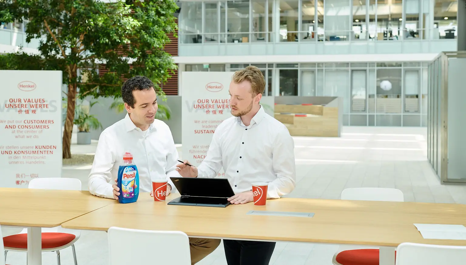 Two men standing at a table with a Persil bottle and a laptop